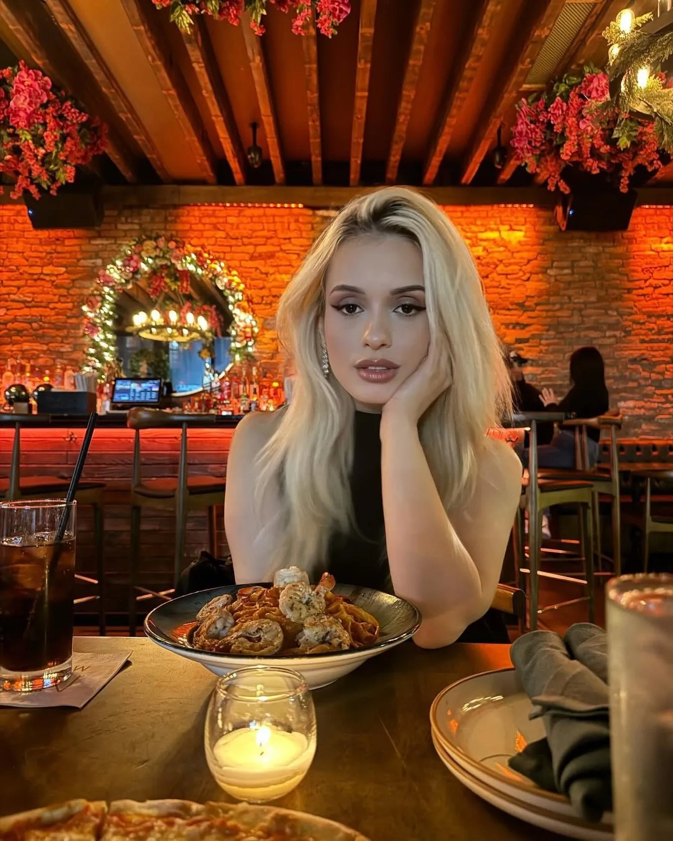 A guest at a candlelit table with a plate of pasta and a pizza, the flower-lined bar behind her.