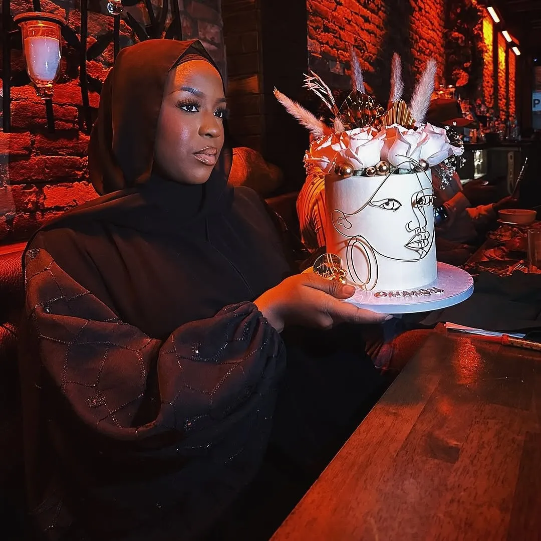 A guest holding a tall white celebration cake with a line-drawn face, lit in warm red.
