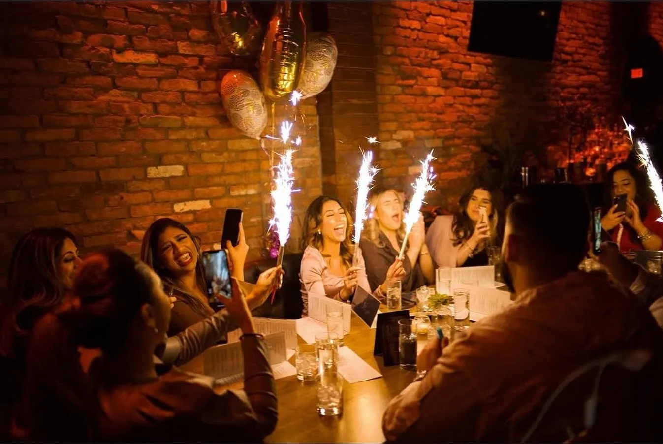 A long table of friends lifting sparklers and phones during a celebration against the brick wall.